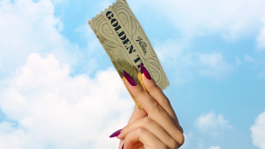 "A hand with long, painted nails holds up a golden ticket against a blue sky with clouds."