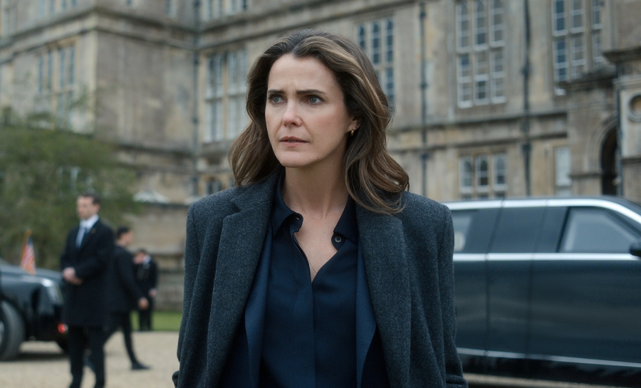 "A woman with wavy brown hair in a dark coat stands outside a grand historic building, looking serious. There is a black car and security personnel in the background, suggesting a formal or official environment."